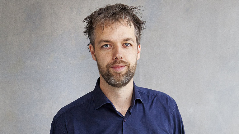 Portret van een man met kort rommelig haar en baard, in een donkerblauw overhemd tegen een grijze achtergrond
