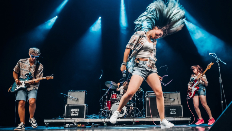 Vijf mensen zitten nonchalant op twee zwarte banken, sommigen met gitaren, gekleed in casual outfits.