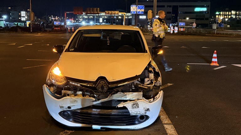 Auto met schade aan de voorkant op een weg 's nachts, met een politieagent en een verkeerskegel in de achtergrond.