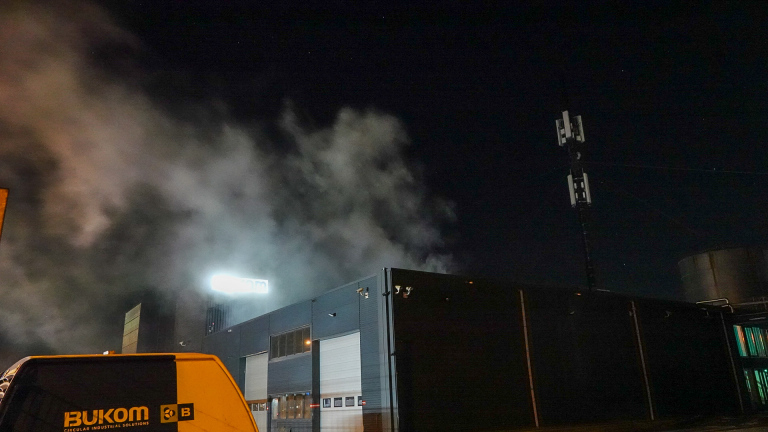 Industrieel gebouw bij nacht, met rookwolken en een zendmast zichtbaar tegen een donkere hemel.