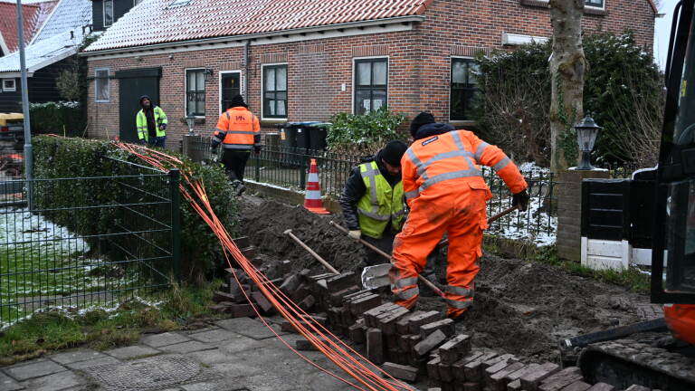 Wegwerkers in oranje en gele kleding graven een greppel langs een trottoir voor een huis.