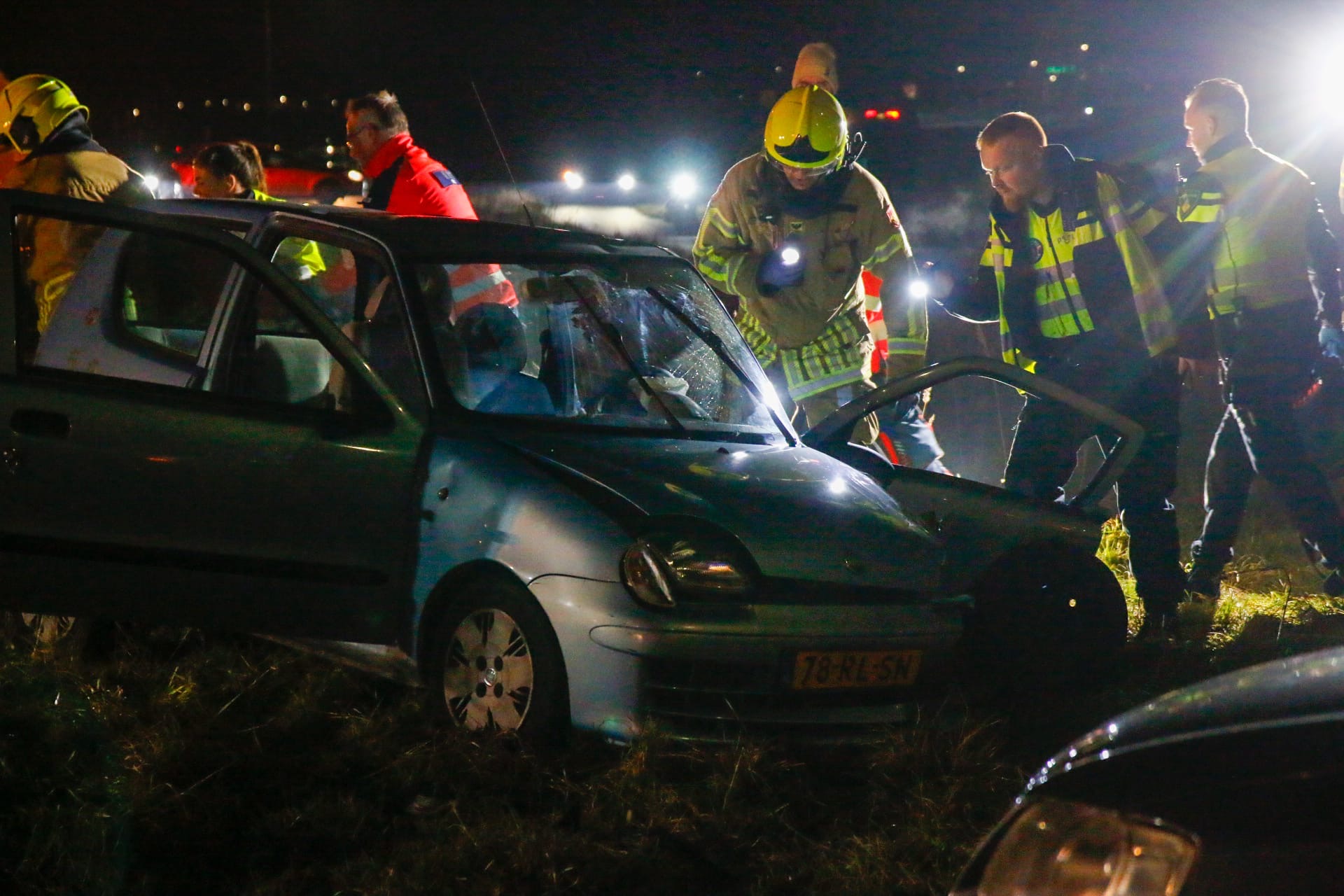 Een blauw beschadigde auto staat langs de weg bij nacht, omringd door hulpverleners en voertuigen met knipperende lichten.