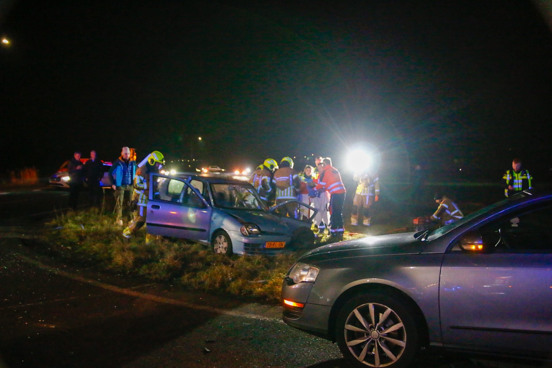 Een blauw beschadigde auto staat langs de weg bij nacht, omringd door hulpverleners en voertuigen met knipperende lichten.