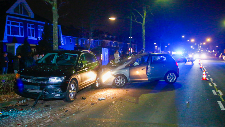 Auto-ongeluk in een woonwijk 's avonds, met twee voertuigen tegen elkaar, glas op de grond en politie-aanwezigheid.