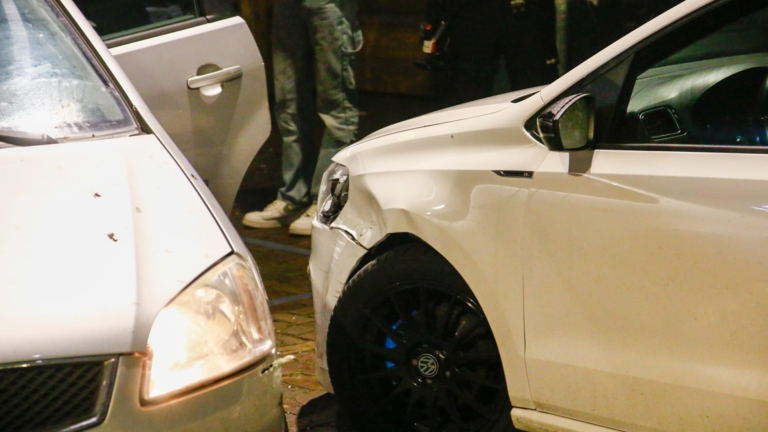 Drie auto's zijn betrokken bij een botsing op een natte straat, met een politieauto en agenten op de achtergrond.