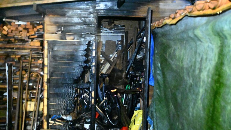 Een schuur met brandschade, gevuld met fietsen en houtblokken, met aan de rechterkant een groene zeilafdekking.