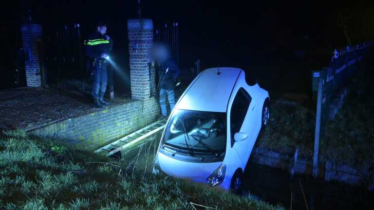 Witte auto rijdt in een sloot bij nacht; politieman en persoon staan bij een stenen muur met de tekst "VEILIGST."