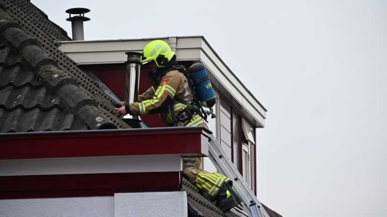 Brandweerman op een ladder controleert een schoorsteen op het dak van een huis terwijl hij beschermende kleding en een helm draagt.
