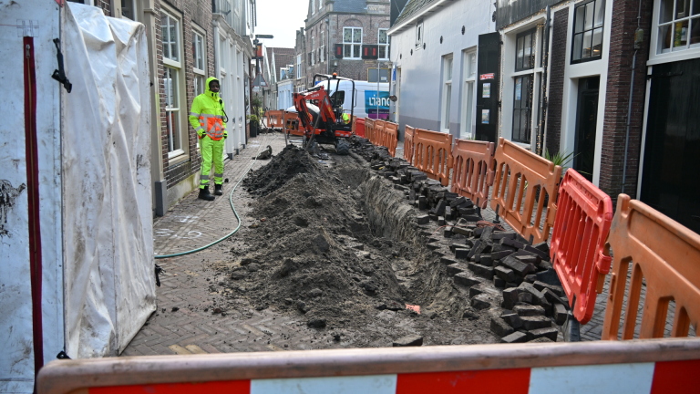 Straat met wegwerkzaamheden, een arbeider in felgeel pak en een graafmachine, omgeven door oranje hekken.