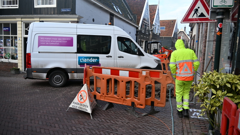 Straat met wegwerkzaamheden, een arbeider in felgeel pak en een graafmachine, omgeven door oranje hekken.