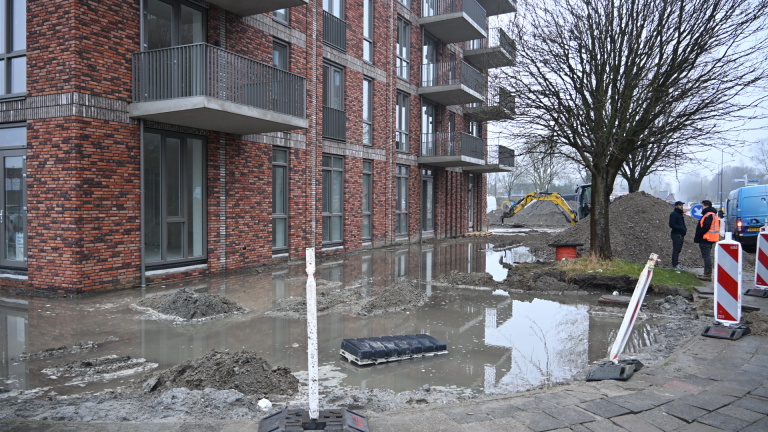 Bouwvakkers werken naast een blauw servicevoertuig rond een modderige waterplas, omgeven door waarschuwingsborden.