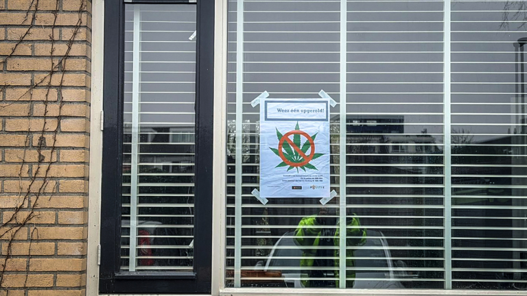 Een huis met een felgroene voordeur en een groot raam met jaloezieën, met een poster op het raam. Een zwarte vuilnisbak staat op de stoep.