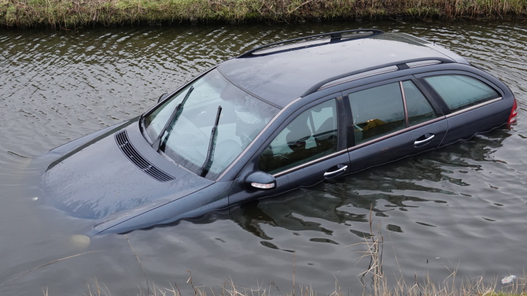 Auto deels ondergedompeld in een sloot langs een weg, met hulpverleners en politievoertuig aanwezig.