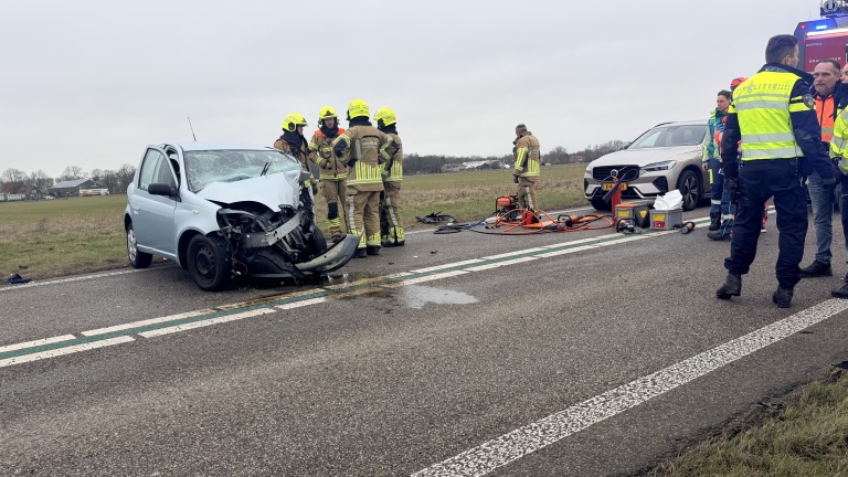 Auto-ongeval op een landelijke weg, met schade aan een kleine blauwe auto en hulpdiensten ter plaatse.