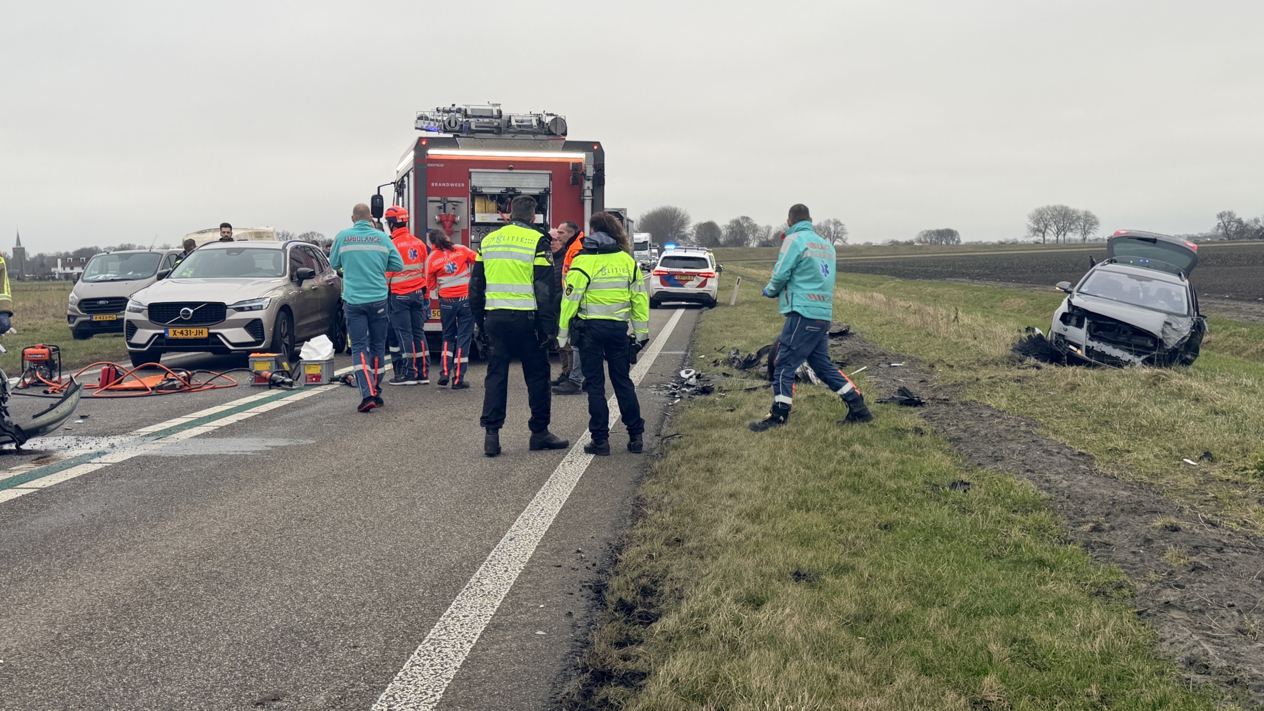 Auto-ongeval op een landelijke weg, met schade aan een kleine blauwe auto en hulpdiensten ter plaatse.