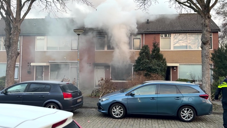 Brandweerauto's staan voor een huis waar dikke rookpluimen uit de ramen komen; een politieagent is aanwezig.
