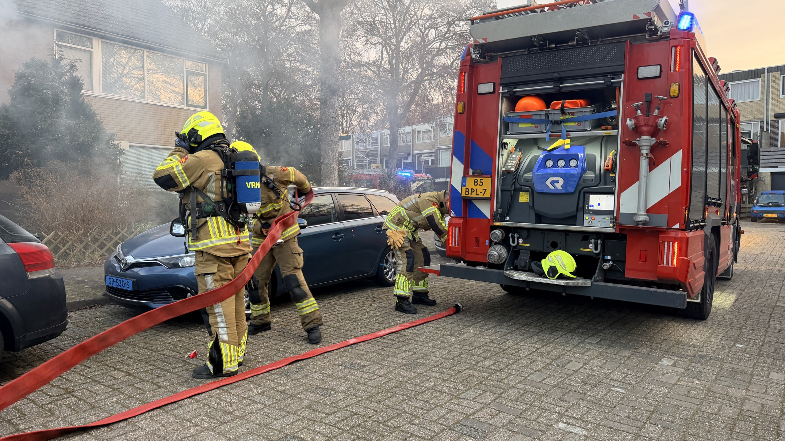 Brandweerauto's staan voor een huis waar dikke rookpluimen uit de ramen komen; een politieagent is aanwezig.