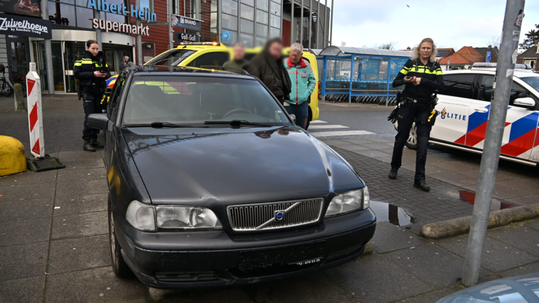 Politieagenten staan bij een geparkeerde auto voor een supermarkt, met een politiewagen op de achtergrond.