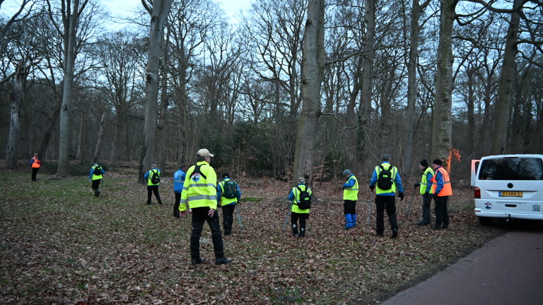 Mensen in gele veiligheidsvesten lopen door een bos met herfstbladeren op de grond, naast een busje.