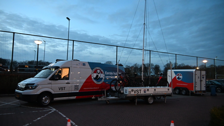 Mensen in gele veiligheidsvesten lopen door een bos met herfstbladeren op de grond, naast een busje.