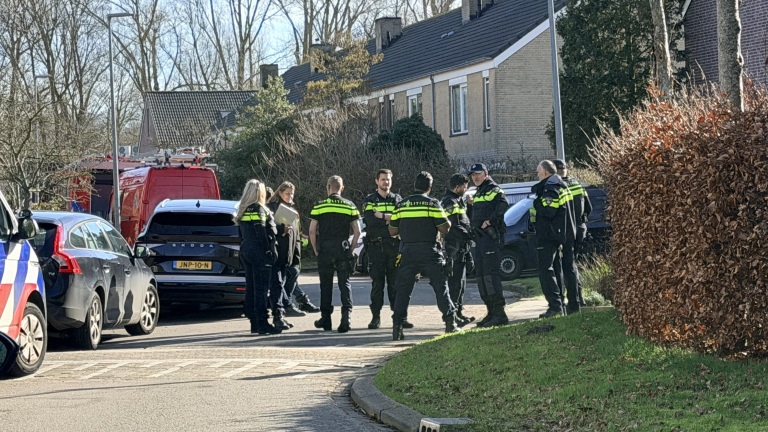 Groep politieagenten in uniform staat buiten bij een woonwijk, naast geparkeerde auto's en een rode wagen.