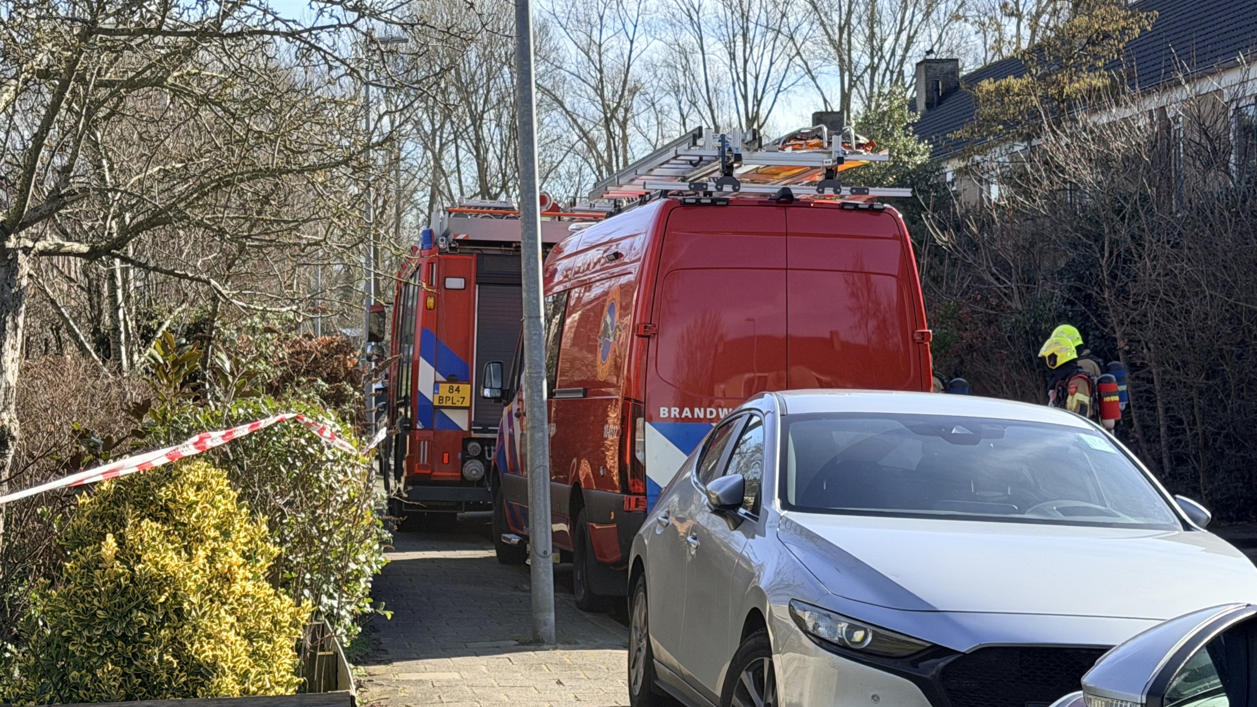 Groep politieagenten in uniform staat buiten bij een woonwijk, naast geparkeerde auto's en een rode wagen.