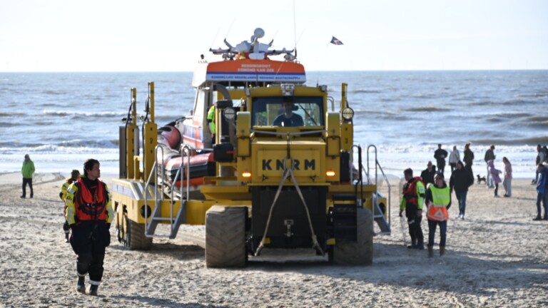 Kusthulpverleningsvoertuig van de KNRM op een strand met personen en een kitesurfer op zee op de achtergrond.