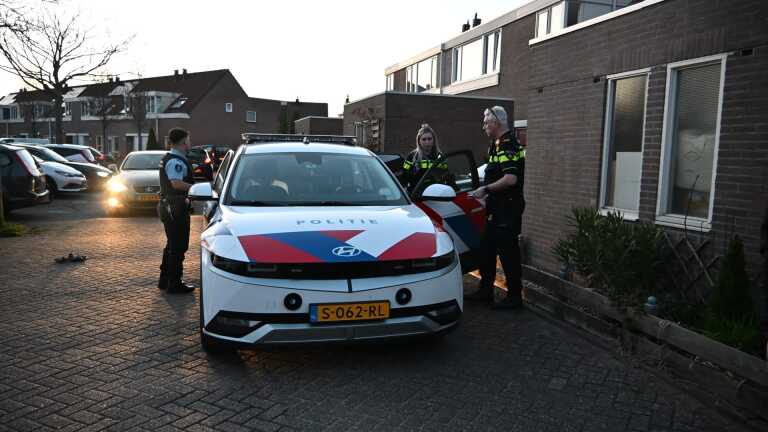 Politieagenten naast een politieauto in een woonwijk, deels verlicht door koplampen van andere auto's.