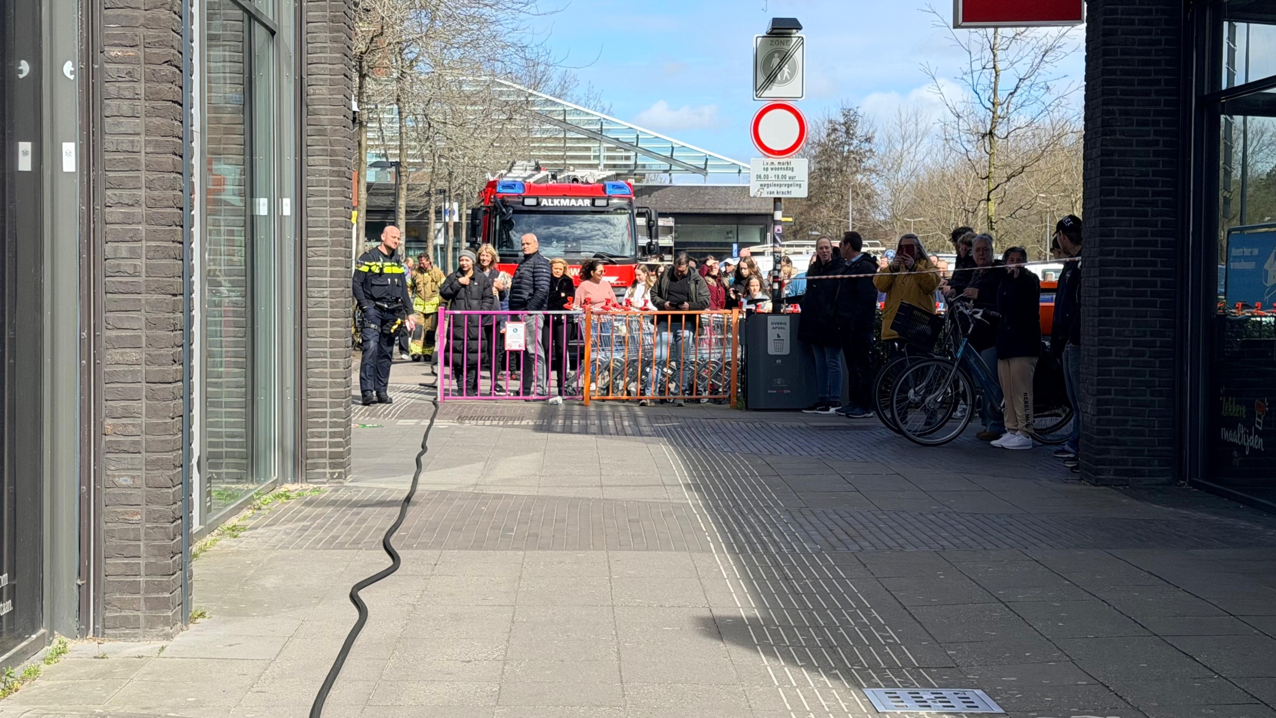 Brandweerlieden en politie bij een winkelstraat, met een politieagent op een motor en Albert Heijn vlaggen zichtbaar.