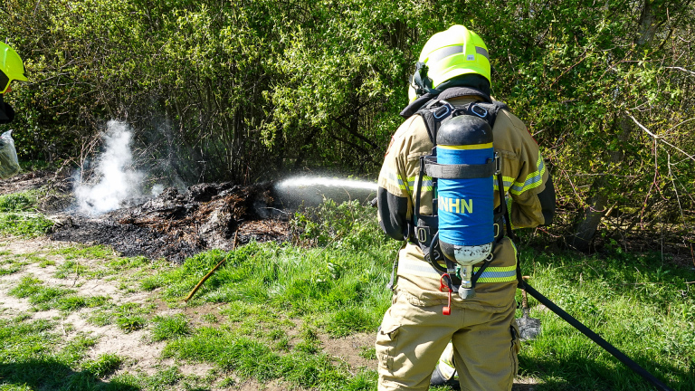 Brandweerman in beschermende kleding blust een smeulend stuk grond naast struiken.
