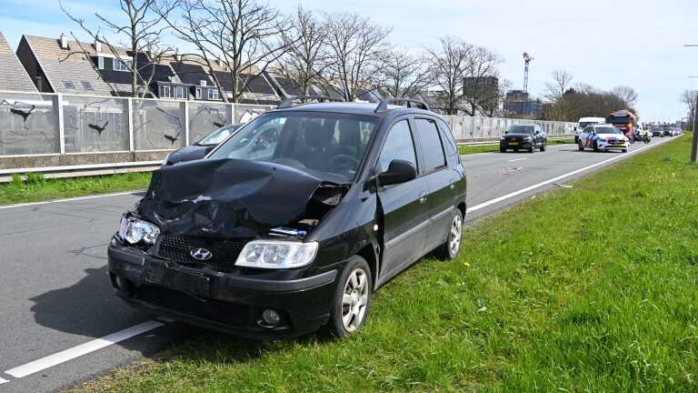 Een zwarte hatchback met een kapotte motorkap staat langs de weg, met politie en verkeer op de achtergrond.