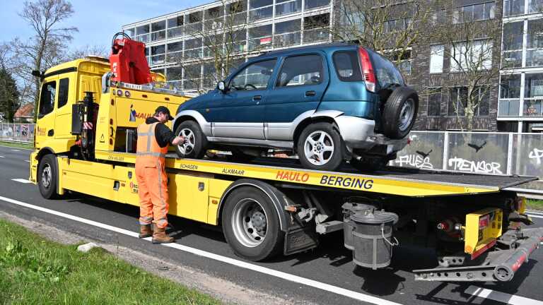 Een zwarte hatchback met een kapotte motorkap staat langs de weg, met politie en verkeer op de achtergrond.