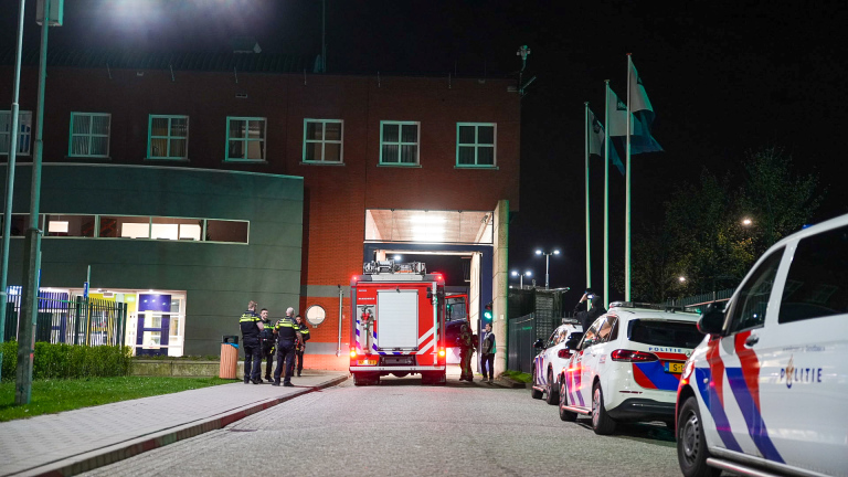 Brandweer- en politievoertuigen met zwaailichten voor een gebouw 's nachts, met zichtbaar personeel bij de ingang.