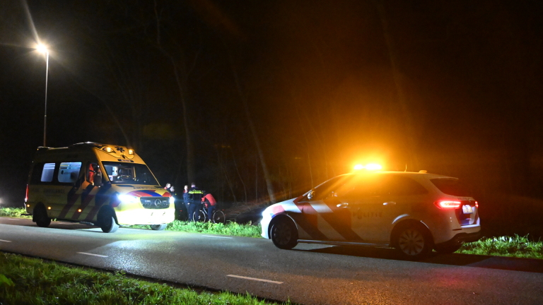 Een ambulance en een politieauto met zwaailichten staan 's nachts langs een weg, omgeven door mensen.