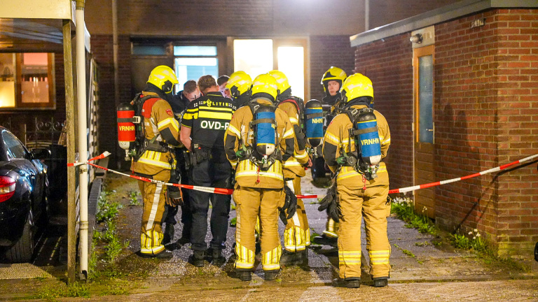 Politieagenten praten buiten een huis terwijl brandweerlieden naar binnen gaan, met een zwarte auto op de oprit.