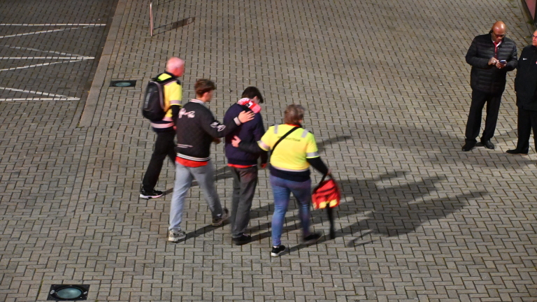 Twee mensen in gele jassen begeleiden een man op een plein, terwijl twee anderen op de achtergrond staan en kijken op hun telefoon.