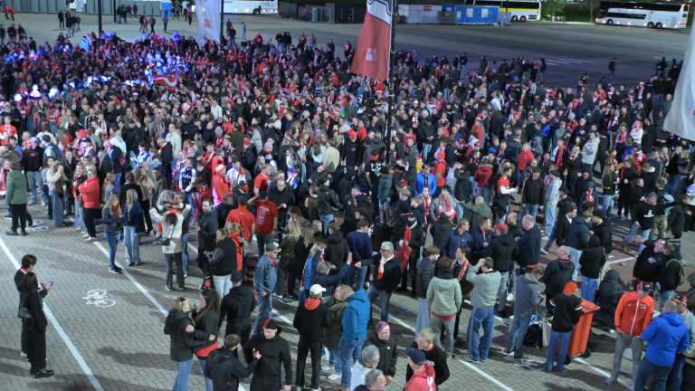 Twee mensen in gele jassen begeleiden een man op een plein, terwijl twee anderen op de achtergrond staan en kijken op hun telefoon.