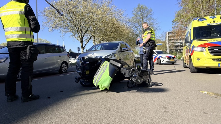Een omgevallen scootmobiel ligt op de weg voor een auto, omringd door politie en een ambulance.