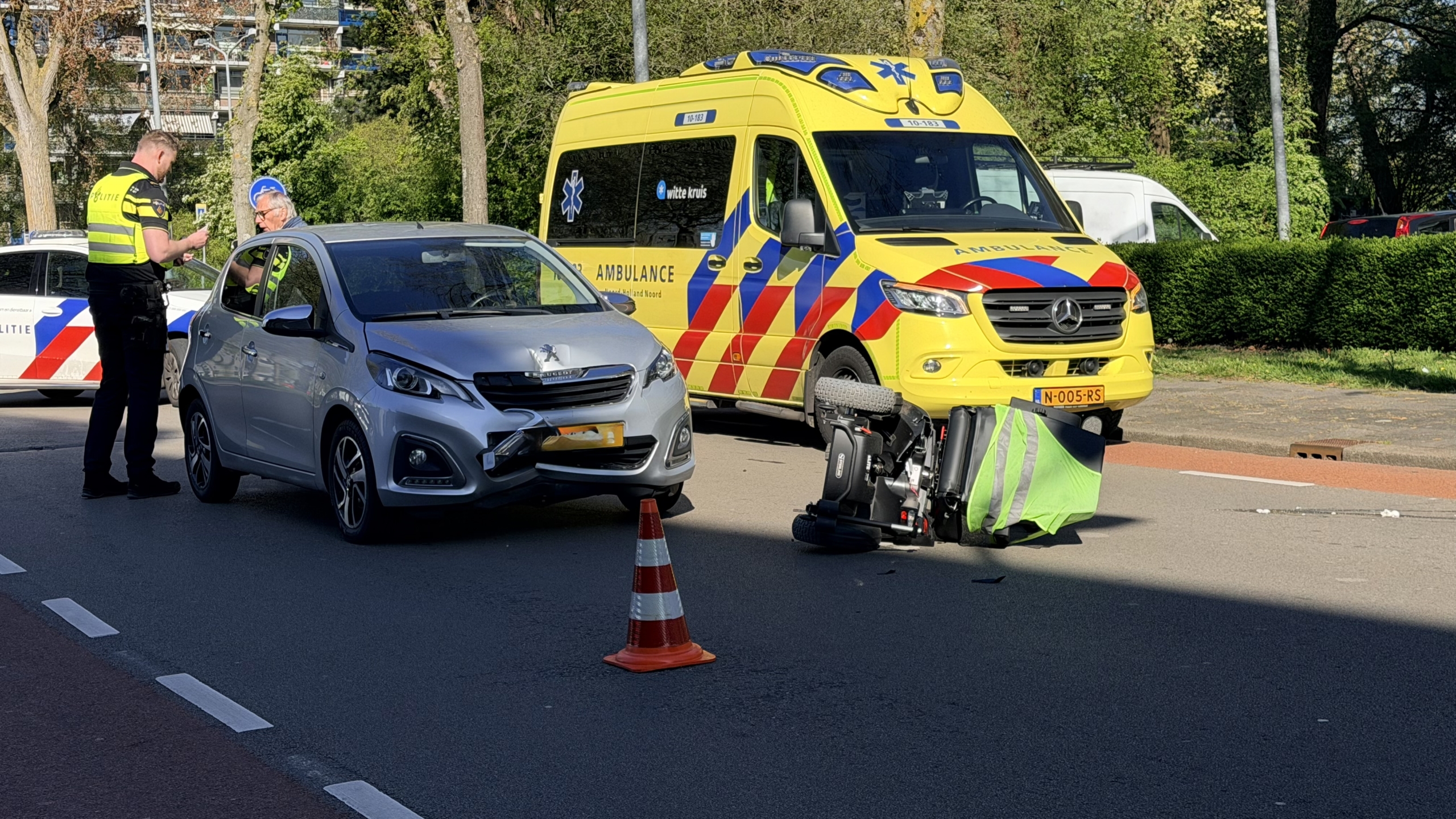 Een omgevallen scootmobiel ligt op de weg voor een auto, omringd door politie en een ambulance.