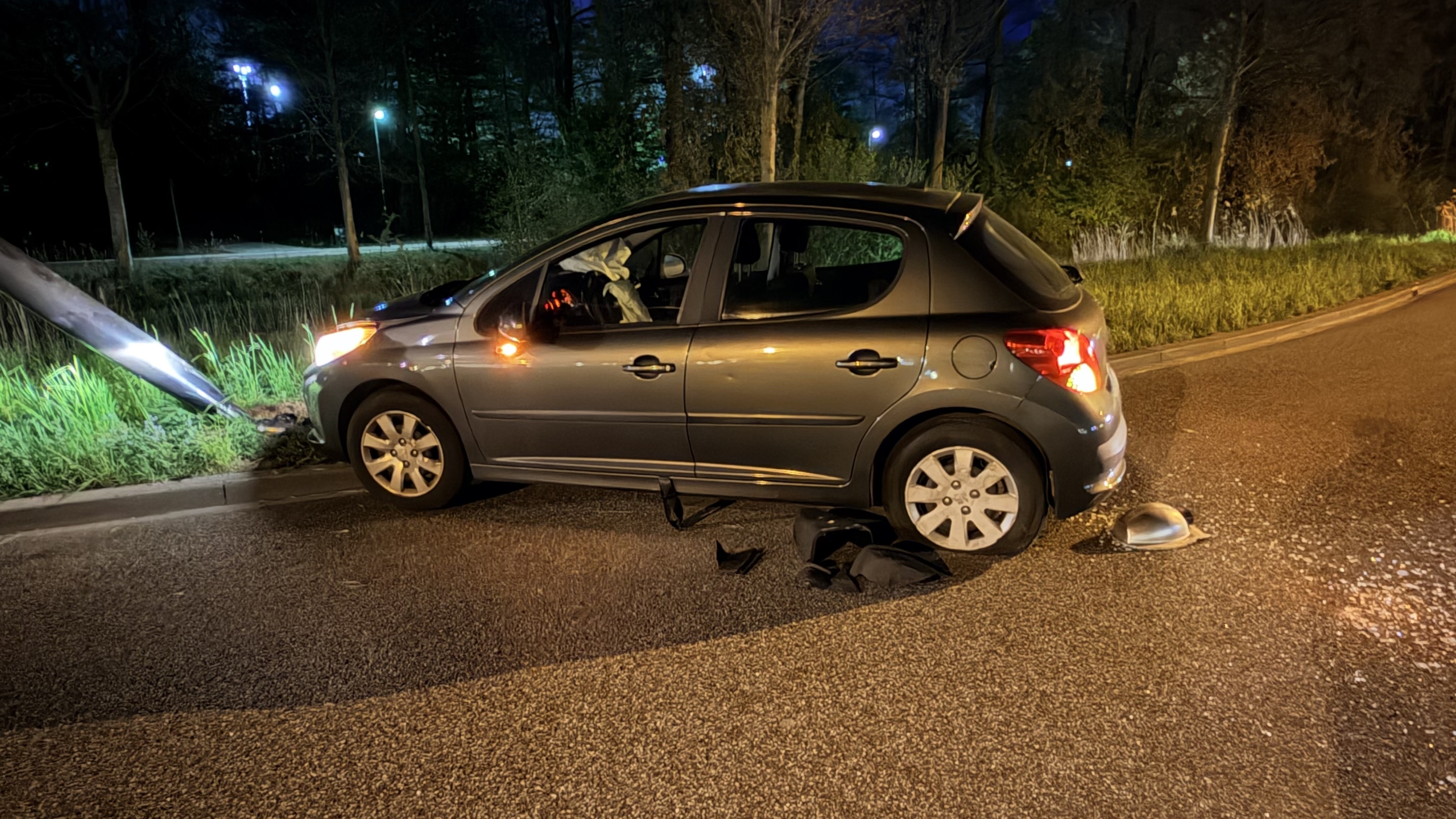 Nachtelijke verkeersscene met een auto die tegen een paal botst, omgeven door hulpverleners en een ambulance, naast een calamiteitenstrookbord.
