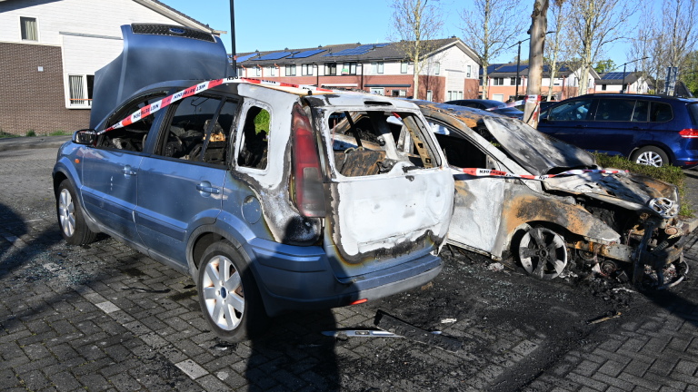 Twee uitgebrande auto's met geopende motorkappen, omgeven door afzetlint, geparkeerd in een woonwijk.