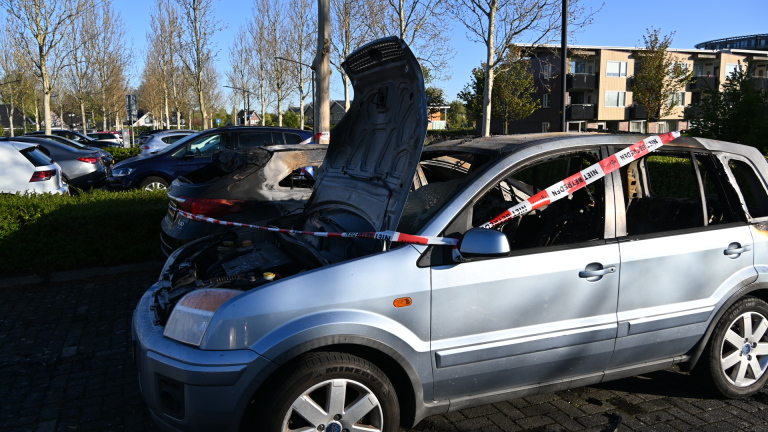 Twee uitgebrande auto's met geopende motorkappen, omgeven door afzetlint, geparkeerd in een woonwijk.