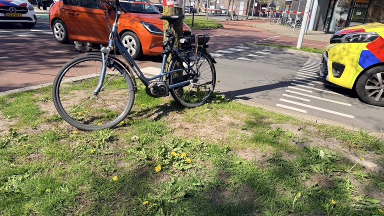 Een fiets is vastgebonden aan een boom naast een weg, naast een felgele auto en een oranje auto.