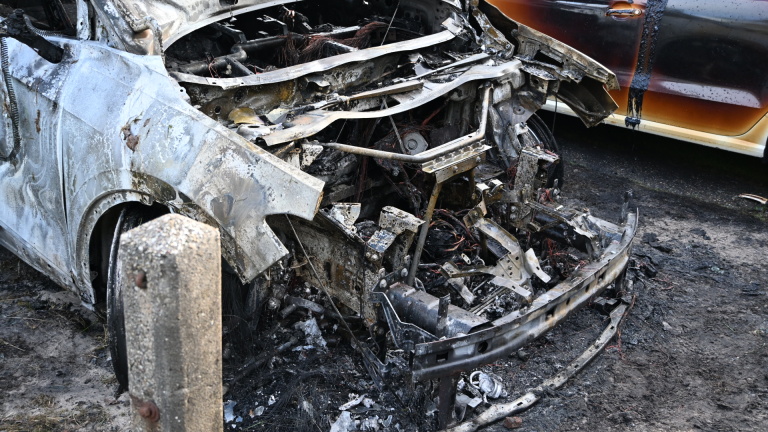 Uitgebrande auto met ernstig beschadigd motorblok en carrosserie, geparkeerd op een onverharde weg.