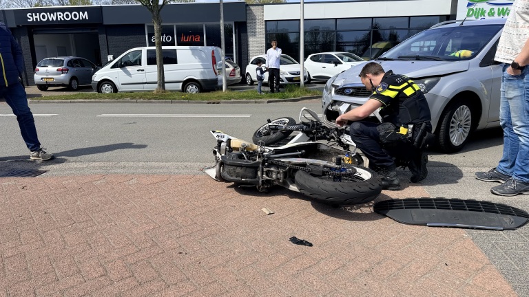 Politieagent onderzoekt een ongeval tussen een motorfiets en een grijze auto, nabij een showroom.