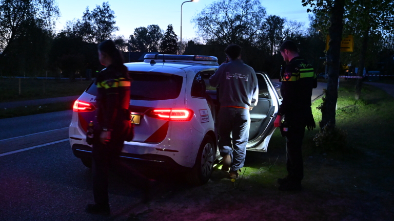 Agenten begeleiden een persoon naar een politieauto in het schemerlicht langs een rustige weg.
