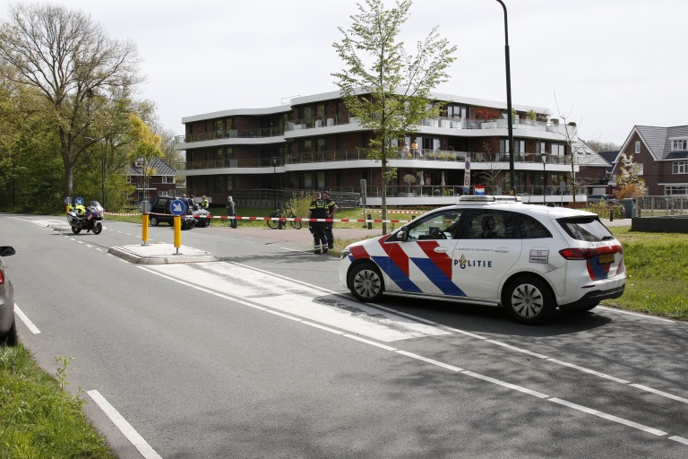 Politievoertuig en motorkap bij afzetting op straat; politieagenten staan bij een groot gebouw.