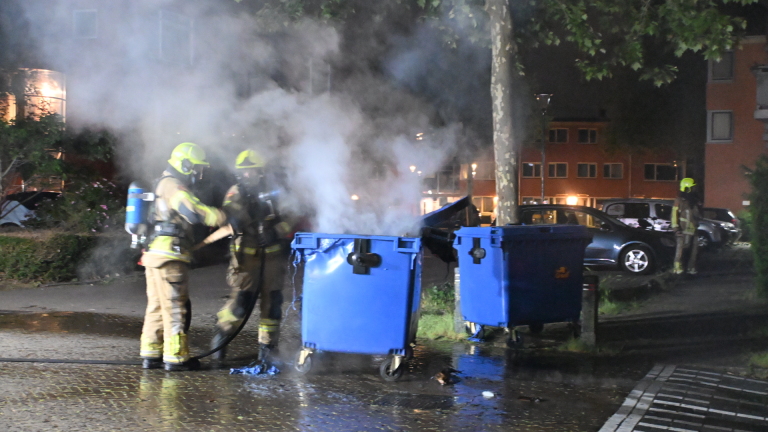 Brandende afvalcontainer naast een straat, met vlammen zichtbaar tegen de nachtelijke achtergrond.