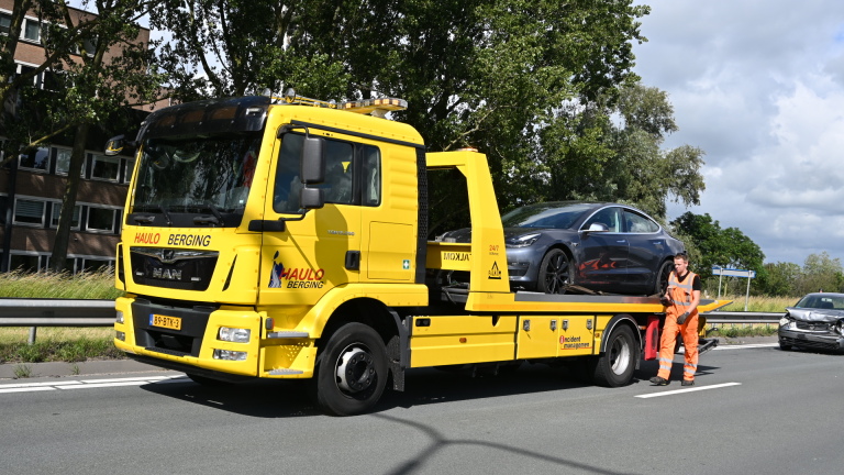 Auto-ongeluk op de snelweg met een beschadigde zilverkleurige auto en een gele bergingswagen die een zwarte auto vervoert.