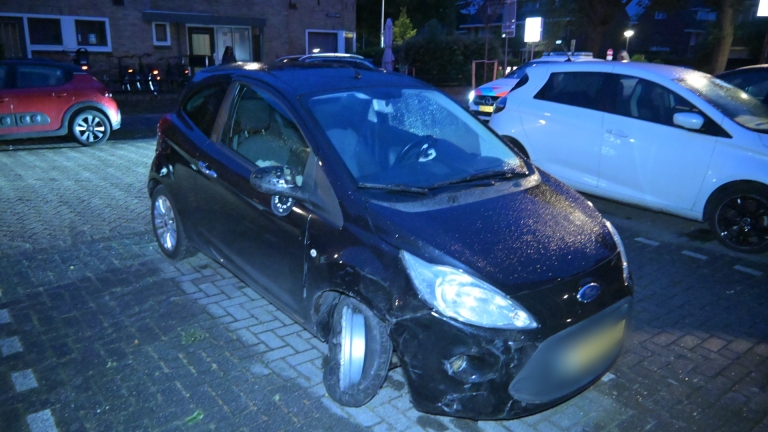Een zwarte auto zonder linkervoorwiel staat beschadigd geparkeerd op een natte straat in het donker.